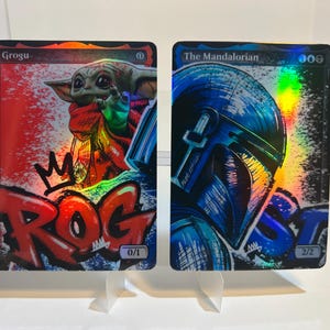 May include: Two collectible cards featuring characters from The Mandalorian. One card shows Grogu with graffiti-style text and a crown. The other card depicts The Mandalorian's helmet in blue tones, with a holographic effect.