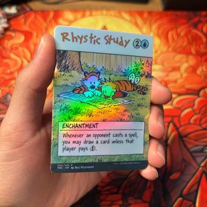 May include: A holographic trading card titled Rhystic Study featuring a colorful illustration of a boy and a tiger reading a newspaper on the grass. The card text reads Enchantment, Whenever an opponent casts a spell, you may draw a card unless that player pays 1.