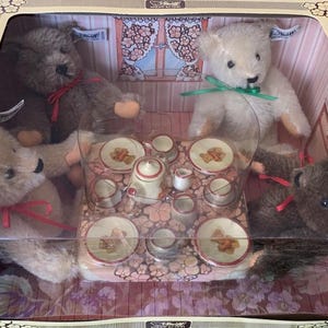 May include: A boxed teddy bear tea party set. The set includes four teddy bears, a miniature tea set with a teapot, cups, and saucers, and a decorative backdrop. The bears are brown and cream-colored, with red and green ribbons.