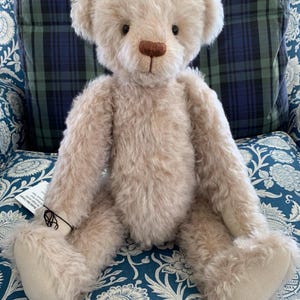 May include: A plush, light brown teddy bear with a soft, furry texture. The bear has a brown nose, dark eyes, and beige paws. It is sitting on a blue floral patterned surface, with a plaid background.