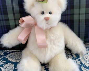 May include: A cream-colored teddy bear with a pink bow and a small pink rose with green leaves on its head. The bear has a fluffy appearance and is sitting on a blue floral patterned surface.