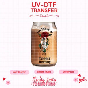 May include: A clear glass can-shaped cup filled with a light brown beverage and ice. The cup features a graphic of a skeleton hand holding a red rose, with the words "you had me at" above and "trigger warning" below.