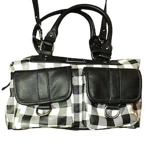 May include: A black and white checkered handbag with two front pockets and black leather accents. The bag has a top handle and a shoulder strap. The pockets have silver D-rings. The bag is a medium size.