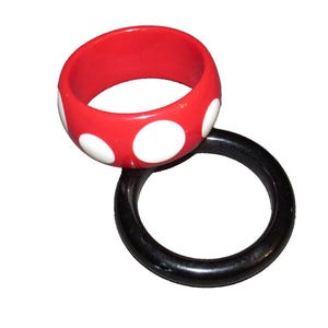 May include: Two bangles. A red, wide bangle with white polka dots is stacked on top of a solid black bangle. The bangles are made of a glossy material and are set against a white background. The red bangle is the focal point.