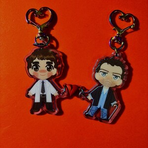 以下が含まれることがあります： 虹色に輝くハート型の留め具が付いた、2つのアクリル製キャラクターキーホルダー。一方は白衣とネクタイを着用し、もう一方はダークジャケットと青いシャツを着て杖を持っています。両者の手の間には小さな赤いハートのチャームが繋がっています。