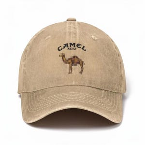 Op de afbeelding: Een beige baseballpet met een verweerde afwerking, voorzien van een afbeelding van een kameel en de tekst CAMEL MIRAGE in het zwart boven het dier gedrukt. De pet heeft een gebogen klep, een knoop aan de bovenkant en gestikte ventilatiegaatjes.