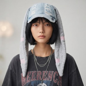 May include: A light blue distressed denim baseball cap featuring long, soft grey fabric bunny ears that hang down on both sides. The grey ears include subtle pink checkered accents. The cap has a structured brim and a worn, frayed texture.
