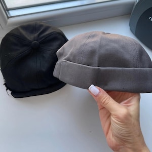 May include: Two hats: a black docker hat with a button on top and a gray docker hat with a folded brim. The gray hat is being held by a hand with manicured nails. Both hats are made of a sturdy fabric.