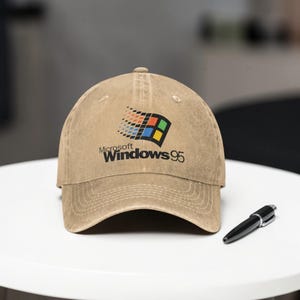 May include: A tan baseball cap featuring the Microsoft Windows 95 logo in the center. The logo is a colorful square design with the text "Microsoft Windows 95" below it. A black pen rests on the white surface.