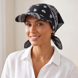 May include: A black headscarf with a white paisley pattern featuring a built-in stiffened brim. The fabric covers the top of the head and ties at the back with two triangular ends, creating a structured visor effect with a classic bandana print.