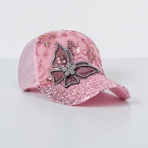 Puede incluir: Gorra de béisbol rosa con parte trasera de malla y visera de lentejuelas. La gorra presenta un gran aplique de mariposa con detalles de pedrería. Bordados florales y lentejuelas decoran la corona, añadiendo brillo.