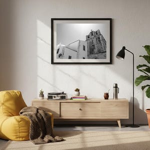 A framed black and white fine art photograph of the Faro Cathedral bell tower hanging in a sunlit modern living room above a light wood console table, next to a cozy yellow beanbag chair and a large potted plant.