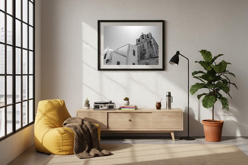 A framed black and white fine art photograph of the Faro Cathedral bell tower hanging in a sunlit modern living room above a light wood console table, next to a cozy yellow beanbag chair and a large potted plant.