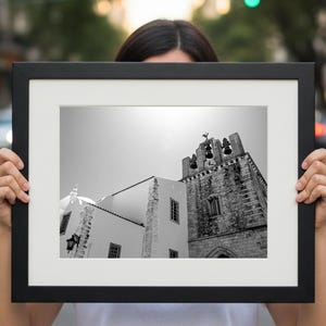 A person standing outdoors holding up a large framed black and white fine art print of a Portuguese church bell tower, with a blurred urban city street in the background.