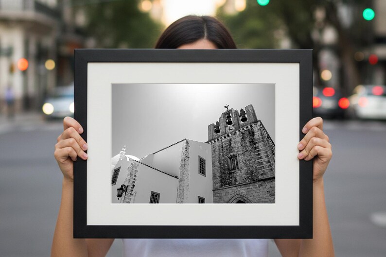 A person standing outdoors holding up a large framed black and white fine art print of a Portuguese church bell tower, with a blurred urban city street in the background.
