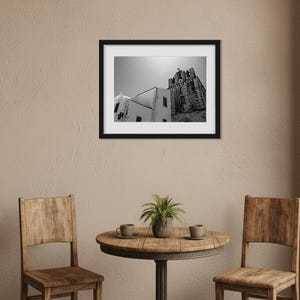 A framed black and white photography print of Faro Cathedral hanging above a small wooden dining table with two chairs, in a cozy rustic room featuring a brick accent wall and a wooden bookshelf.