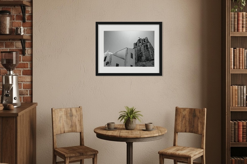 A framed black and white photography print of Faro Cathedral hanging above a small wooden dining table with two chairs, in a cozy rustic room featuring a brick accent wall and a wooden bookshelf.