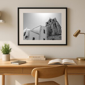 A framed black and white architectural photograph of a historic bell tower in Portugal, hanging on a sun-drenched wall above a modern minimalist wooden desk with an open book, chair, and a gold lamp