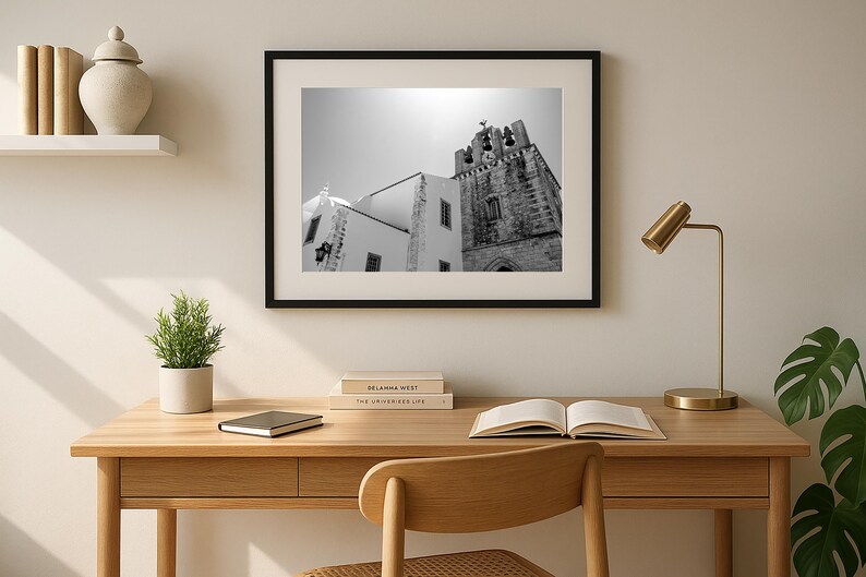 A framed black and white architectural photograph of a historic bell tower in Portugal, hanging on a sun-drenched wall above a modern minimalist wooden desk with an open book, chair, and a gold lamp