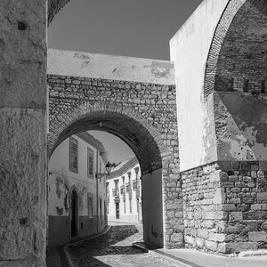 Black and white fine art photography of a medieval stone archway in Faro Old Town Portugal. Vertical architecture print showing rustic cobblestone street details and historic city entrance.