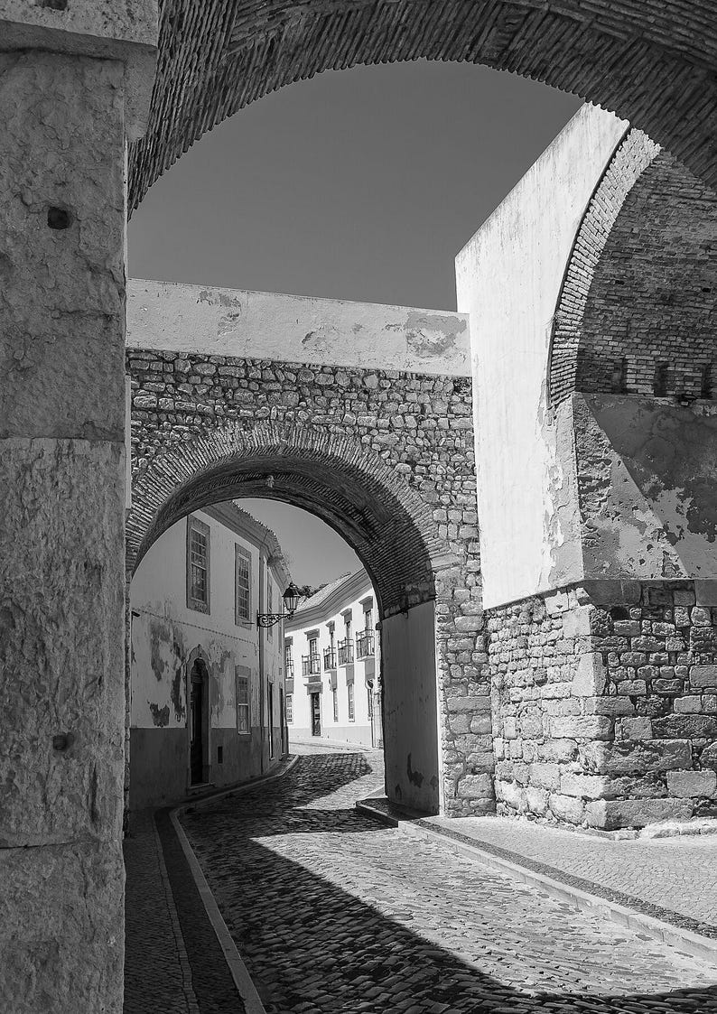 Black and white fine art photography of a medieval stone archway in Faro Old Town Portugal. Vertical architecture print showing rustic cobblestone street details and historic city entrance.