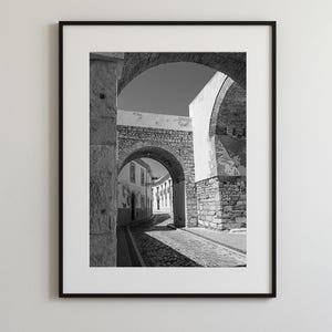 Black and white framed photography of a medieval stone archway in Faro Old Town Portugal. Historic cobblestone street architecture wall art with a white mat.
