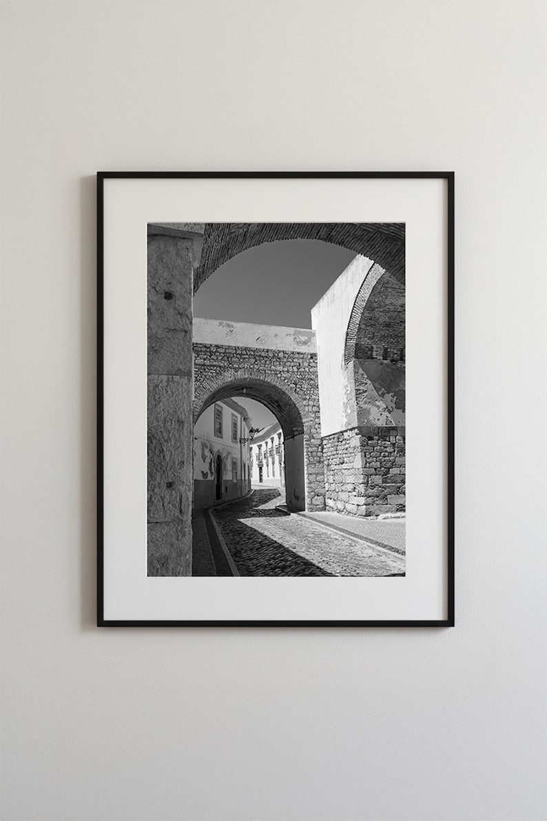 Black and white framed photography of a medieval stone archway in Faro Old Town Portugal. Historic cobblestone street architecture wall art with a white mat.