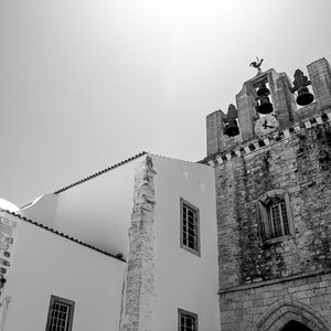 A striking black and white architectural photograph of the ancient stone bell tower of Faro Cathedral in Portugal, contrasting against a bright white building and a clear summer sky.