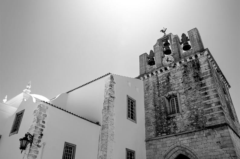 A striking black and white architectural photograph of the ancient stone bell tower of Faro Cathedral in Portugal, contrasting against a bright white building and a clear summer sky.
