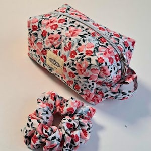 May include: A floral print cosmetic bag and scrunchie set. The bag is rectangular with a zipper closure and a small handle. The fabric features a pattern of red and pink flowers on a white background. The scrunchie matches the bag's floral design.