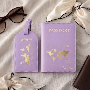 Könnte beinhalten: Ein lavendelfarbenes Reisepassetui und ein Gepäckanhänger-Set mit goldenen Weltkartenmustern und dem Namen "Maria". Auf dem Reisepassetui steht "PASSPORT". Sonnenbrille und Schal im Hintergrund.