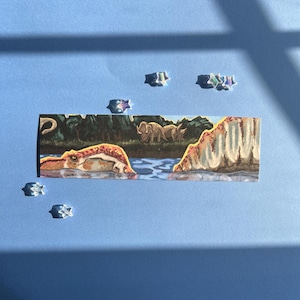 May include: A rectangular bookmark featuring a painted scene of a prehistoric landscape with a river, a large dinosaur head emerging from the water, and triceratops in the background. The bookmark is surrounded by several small, clear star-shaped accents.