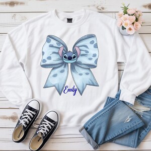 Puede incluir: Sudadera blanca con un lazo azul claro con un personaje de dibujos animados y el nombre "Emily". El lazo tiene un estampado de lunares. Zapatillas de lona negras y vaqueros azules tambi&eacute;n en la imagen.