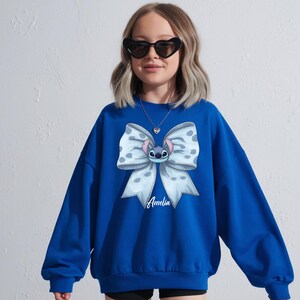 Puede incluir: Una sudadera azul real con un gran dise&ntilde;o de lazo y un personaje de dibujos animados en el centro. El lazo es blanco con lunares azules. El nombre "Amelia" est&aacute; escrito en cursiva debajo. La persona lleva gafas de sol en forma de coraz&oacute;n.