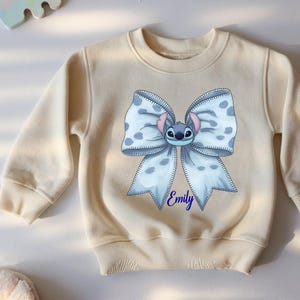 Puede incluir: Sudadera de color crema con un gran dise&ntilde;o de lazo con la cara de un personaje de dibujos animados en el centro. El lazo es azul claro con lunares blancos. El nombre "Emily" est&aacute; escrito en azul debajo.