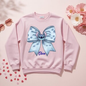 Puede incluir: Sudadera rosa claro con un gran lazo azul con lunares blancos y un personaje de dibujos animados en el centro. El nombre "Olivia" est&aacute; escrito en azul debajo. La sudadera tiene mangas largas.