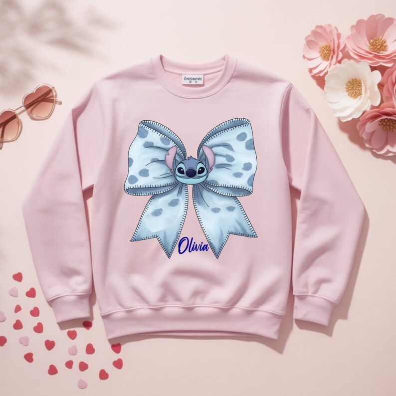 Puede incluir: Sudadera rosa claro con un gran lazo azul con lunares blancos y un personaje de dibujos animados en el centro. El nombre "Olivia" est&aacute; escrito en azul debajo. La sudadera tiene mangas largas.