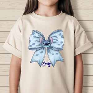 Puede incluir: Camiseta beige claro con un gran lazo azul con lunares blancos y una cara de personaje de dibujos animados. El nombre "Emily" est&aacute; escrito en azul debajo del lazo. La camiseta tiene mangas cortas.