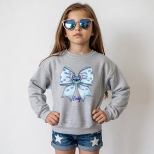Puede incluir: Una sudadera gris con un lazo azul claro que presenta un personaje de dibujos animados y el nombre "Emily" en azul. El lazo tiene un estampado de lunares. La persona lleva gafas de sol en forma de coraz&oacute;n y pantalones cortos vaqueros.
