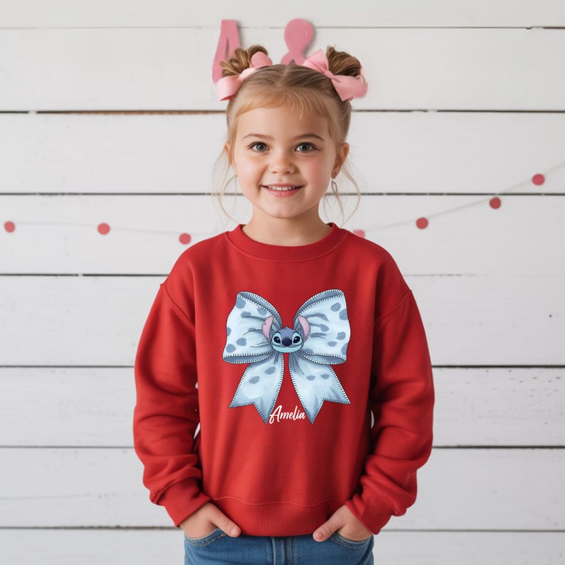 Puede incluir: Una sudadera roja con un gran lazo azul claro con la cara de un personaje de dibujos animados y el nombre "Amelia" en escritura blanca. El lazo tiene un estampado de lunares. Llevada por una ni&ntilde;a.