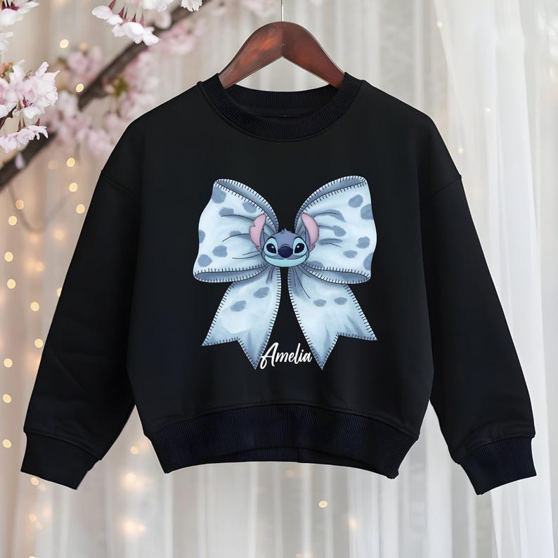 Puede incluir: Sudadera negra con un lazo azul claro grande con lunares blancos y un personaje de dibujos animados en el centro. El nombre "Amelia" est&aacute; escrito debajo. La sudadera est&aacute; colgada en una percha de madera.