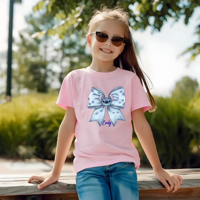 Puede incluir: Camiseta rosa con un lazo azul claro con lunares blancos y un personaje de dibujos animados en el centro. El nombre "Emily" est&aacute; impreso en azul debajo. La camiseta la lleva una persona joven.