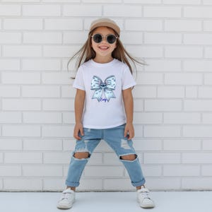 Puede incluir: Camiseta blanca con un dise&ntilde;o de lazo azul, un personaje de dibujos animados y el nombre "Emily". El conjunto incluye vaqueros rotos azul claro y zapatillas blancas. La persona lleva una gorra beige y gafas de sol.
