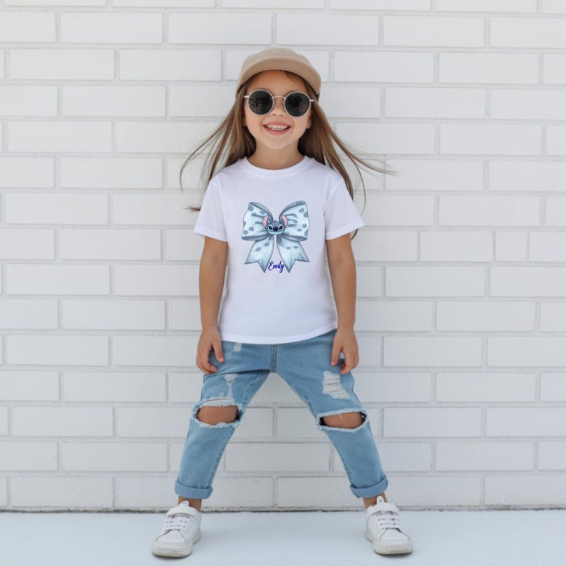Puede incluir: Camiseta blanca con un dise&ntilde;o de lazo azul, un personaje de dibujos animados y el nombre "Emily". El conjunto incluye vaqueros rotos azul claro y zapatillas blancas. La persona lleva una gorra beige y gafas de sol.