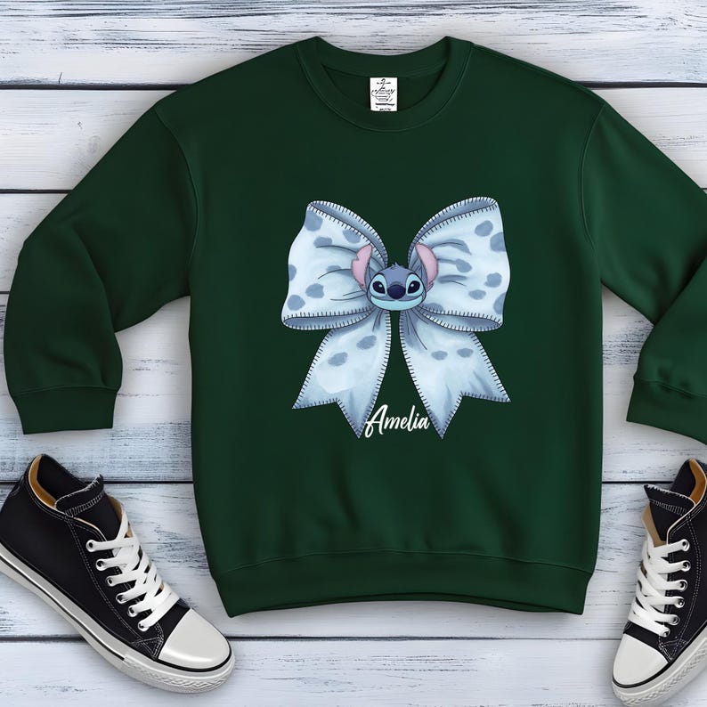 Puede incluir: Una sudadera verde oscuro con un lazo azul claro con lunares blancos y un personaje de dibujos animados. El nombre "Amelia" est&aacute; impreso en cursiva en el lazo. Zapatillas deportivas blancas y negras son visibles en primer plano.