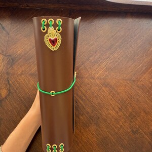 May include: A brown leather journal with gold-colored accents and a green elastic band. The journal features a decorative gold heart with a red center and is held upright.