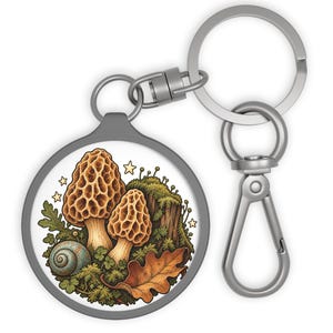 May include: A circular keychain featuring a silver-toned metal ring and clasp. The central disc displays a detailed illustration of two tan morel mushrooms, a small blue snail, a brown oak leaf, and green moss, accented by several small yellow stars.