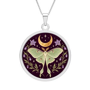 May include: A silver-toned circular pendant necklace featuring a central illustration of a pale green moth with long wings. The moth is framed by purple bell-shaped flowers against a dark purple background, topped with a golden crescent moon.
