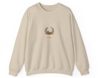 Op de afbeelding: Een beige sweater met ronde hals en een kleine, gecentreerde afbeelding van een krans van eikels en eikenbladeren. Onder de krans staat de naam Eleanor gedrukt in een donker schreeflettertype. Lange mouwen en een geribbelde hals.