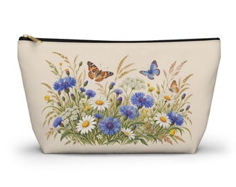 May include: A beige zipped pouch featuring a botanical illustration of blue cornflowers, white daisies, and tall grasses. Three butterflies in shades of orange and blue are depicted hovering above the floral arrangement.